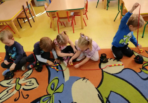 Dzieci siedzą na dywanie i nawlekają klocki numicon na sznurki według właściwej kolejności.