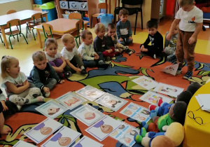 Dzieci siedzą na dywanie, na dywanie rozłożone są obrazki przedstawiające róźne emocje, na dywanie siedzą również misie, chłopiec stoi na dywanie.