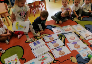 Dzieci siedzą na dywanie, na dywanie rozłożone są obrazki przedstawiające róźne emocje, na dywanie siedzą również misie, dziewczynka stoi na dywanie i trzyma jeden obrazek.