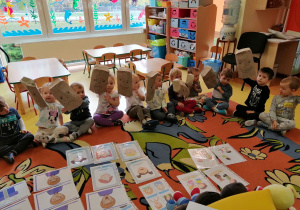 Dzieci siedzą na dywanie, trzymają w rękach papierowe torby, na których nrysowane są różne emocje.