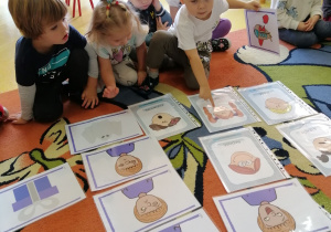 Dzieci siedzą na dywanie, na dywanie rozłożone są obrazki przedstawiające róźne emocje, chłopiec wskazuje jeden obrazek, a drugi trzyma w ręce.