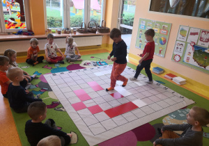 Dzieci siedzą na dywanie, dwóch chłopców odkodowuje miejsca tabliczek na macie do kodowania.