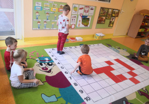Dzieci siedzą na dywanie, dwóch chłopców odkodowuje miejsca tabliczek na macie do kodowania.