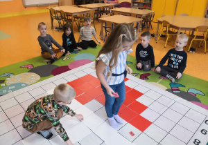 Dzieci siedzą na dywanie, dziewczynka i chłopiec odkodowują miejsca tabliczek na macie do kodowania.