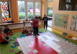 Dzieci siedzą na dywanie, trzech chłopców odkodowuje miejsca tabliczek na macie do kodowania.