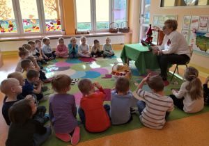 Przedszkolaki siedzą na dywanie i słuchają bajki czytanej przez Panią Marię.