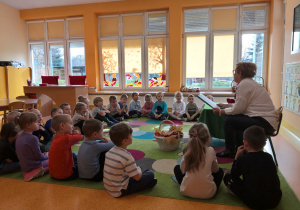 Przedszkolaki siedzą na dywanie i słuchają bajki czytanej przez Panią Marię.