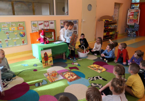 Przedszkolaki siedzą na dywanie, chłopiec pokazuje dzieciom ilustracje związaną z czytaną bajką.