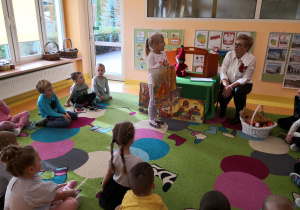 Przedszkolaki siedzą na dywanie, dziewczynka pokazuje dzieciom ilustracje związaną z czytaną bajką.
