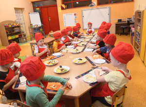 Dzieci siedzą przy stolikach, są ubrani w fartuszki i czapki kucharskie, kroją owoce i nadziewają je na patyczki do szaszłyków.