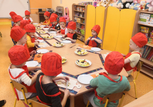 Dzieci siedzą przy stolikach, są ubrani w fartuszki i czapki kucharskie, nadziewają pokrojone owoce na patyczki do szaszłyków.