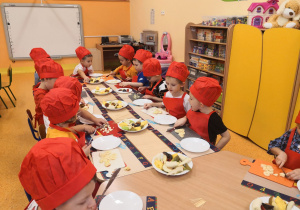 Dzieci siedzą przy stolikach, są ubrani w fartuszki i czapki kucharskie, kroją owoce.