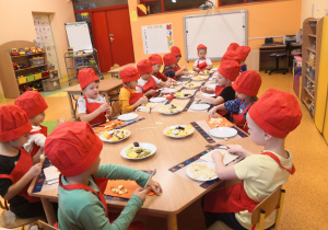 Dzieci siedzą przy stolikach, są ubrani w fartuszki i czapki kucharskie, kroją owoce i nadziewają je na patyczki do szaszłyków.