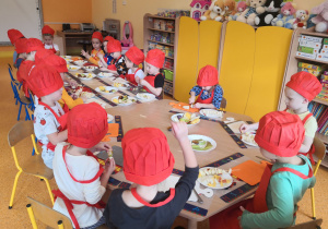 Dzieci siedzą przy stolikach, są ubrani w fartuszki i czapki kucharskie, kroją owoce i nadziewają je na patyczki do szaszłyków.