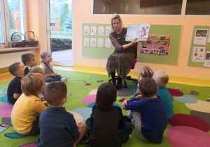 Przedszkolaki siedzą na dywanie, mama Leona pokazuje im ilustracje z bajki.