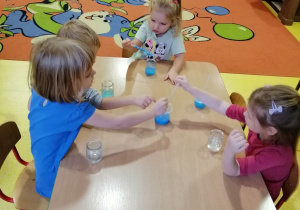 Dzieci siedzą przy stoliku i nabierają barwnik do pipetek.