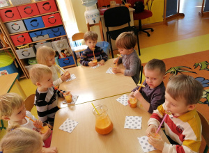 Dzieci siedzą przy stolikach i piją sok przez słomki.