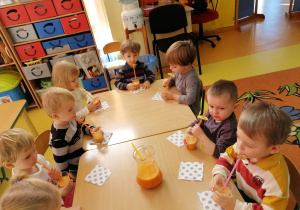 Dzieci siedzą przy stolikach i piją sok przez słomki.