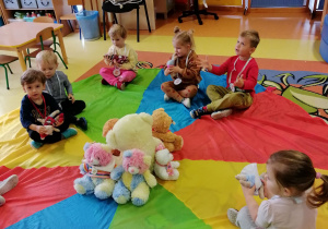 Dzieci siedzą na chuście animacyjnej, na środku chusty siedzą misie.
