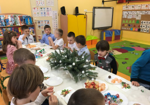 Dzieci siedzą przy wigilijnym stole i jedzą pyszne słodkości oraz owoce.