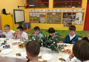 Dzieci siedzą przy wigilijnym stole i jedzą pyszne słodkości oraz owoce.