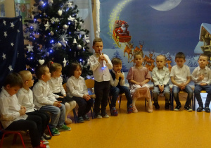 Dzieci siedzą, chłopiec stoi trzyma mikrofon i mówi wiersz.
