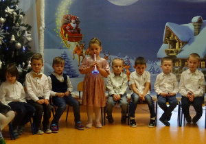 Dzieci siedzą, dziewczynka stoi trzyma mikrofon i mówi wiersz.