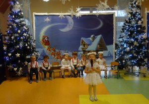 Dzieci siedzą, dziewczynka stoi na środku holu i recytuje wiersz.