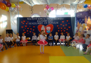 Dzieci siedzą na krzesłach, dziewczynka stoi na środku holu i recytuje swój wiersz.