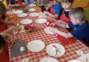 Dzieci siedzą przy stolikach i wykonują wielkanocne zajączki z warzyw i jajek.