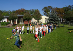 Przedszkolaki są w ogrodzie przedszkolnym, są ustawieni w dwóch rzędach, chłopiec biegnie z piłką na paletce.