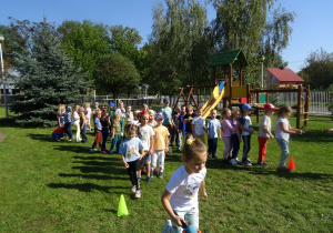 Przedszkolaki są w ogrodzie przedszkolnym, są ustawieni w dwóch rzędach, dziewczynka biegnie z piłką na paletce.