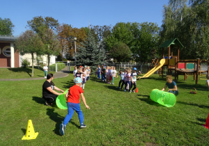 Przedszkolaki są w ogrodzie przedszkolnym, na zdjęciu jest Pani nauczycielka i Pani woźna, które trzymają tunele, dzieci są ustawione w dwóch rzędach, chłopiec przeszedł właśnie przez tunel.