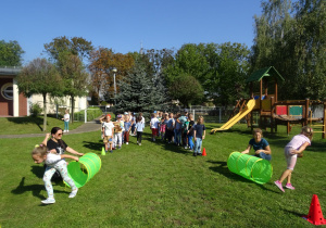 Przedszkolaki są w ogrodzie przedszkolnym, na zdjęciu jest Pani nauczycielka i Pani woźna, które trzymają tunele, dzieci są ustawione w dwóch rzędach, dwie dziewczynki z konkurencyjnych drużyn przeszły właśnie przez tunele.