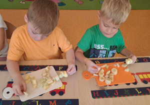 Chłopcy siedzą przy stoliku, na stoliku leżą podkładki oraz deski, chłopcy nadziewają owoce na patyczki.